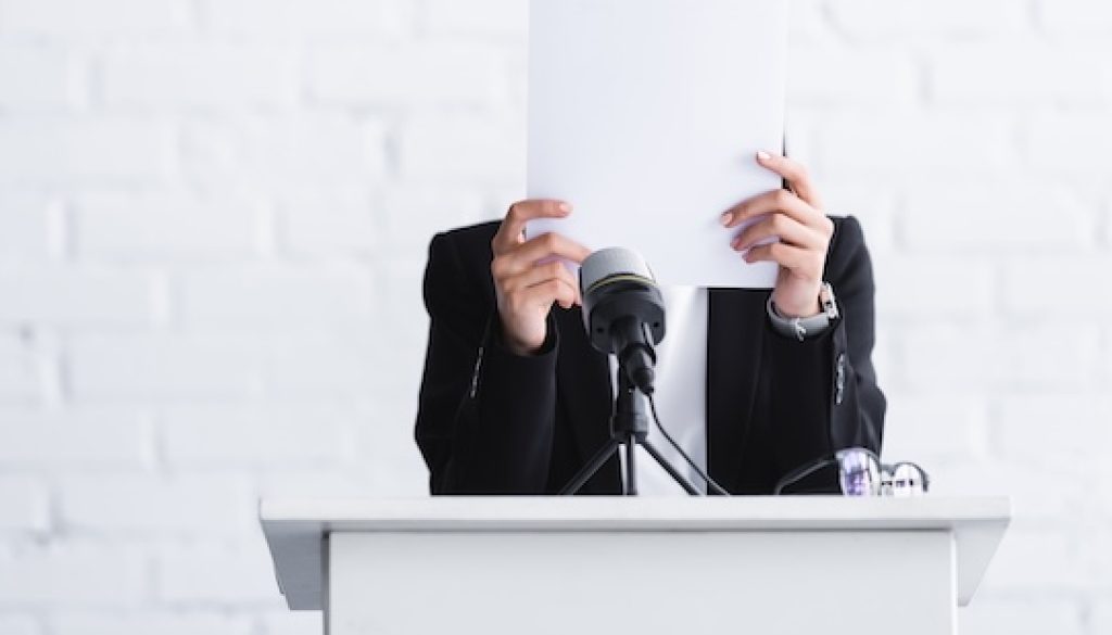 young lecturer, suffering from fear of public speaking, standing on podium tribune and hiding face